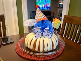 A bundt cake with cream drizzle is adorned with decorations for a 56th birthday celebration. A colorful birthday hat, blue tissue paper decoration, and a 'Happy Bundtday' sign enhance the festive atmosphere. The cake sits on a round red placemat on a wooden table beside a smartphone. In the background, a television displays a sports game.