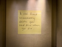 A handwritten note on a yellow sticky note is placed on a textured wall. The note humorously states that a true friend would delete your hard drive immediately after you die.