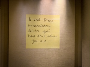 A handwritten note on a yellow sticky note is placed on a textured wall. The note humorously states that a true friend would delete your hard drive immediately after you die.