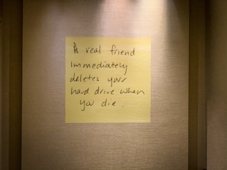 A handwritten note on a yellow sticky note is placed on a textured wall. The note humorously states that a true friend would delete your hard drive immediately after you die.