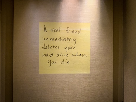 A handwritten note on a yellow sticky note is placed on a textured wall. The note humorously states that a true friend would delete your hard drive immediately after you die.