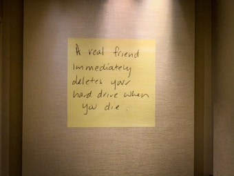 A handwritten note on a yellow sticky note is placed on a textured wall. The note humorously states that a true friend would delete your hard drive immediately after you die.