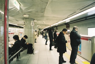 A bustling subway platform with a diverse group of people waiting for the train. Some are seated while others stand, engaged with their mobile phones. The station is well-lit, and a train is visible on the opposite track.