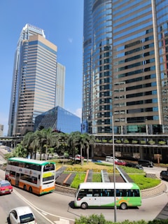 A bustling urban scene featuring modern skyscrapers with glass facades. The area includes a circular road with a green roundabout featuring palm trees and flower beds. Several vehicles, including a double-decker bus and cars, navigate the road. The sky is clear and blue, enhancing the sleek architectural lines of the buildings.