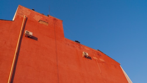A red building facade with visible patches of exposed brickwork, featuring two air conditioning units mounted on the wall. A vertical pipe and an antenna can also be seen. The building contrasts against a clear blue sky.