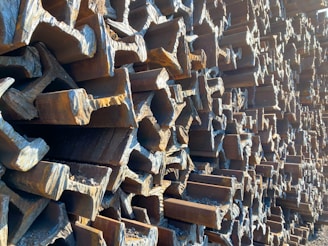 A large pile of rusty, weathered steel beams stacked in an interlocking pattern creating a complex, geometric texture. The light casts shadows, enhancing the rough surfaces and edges of the beams.