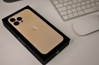 A new golden iPhone in its black box is placed on a white table alongside a white keyboard and mouse. The glossy apple logo is visible on the phone and box, with the phone’s triple-camera setup prominently displayed.