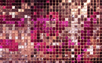 A close-up view of a shiny mosaic wall composed of numerous square panels. The panels are arranged in a grid pattern and vary in shades of pink, gold, and rose gold. The reflective surface of the panels creates a shimmering effect.