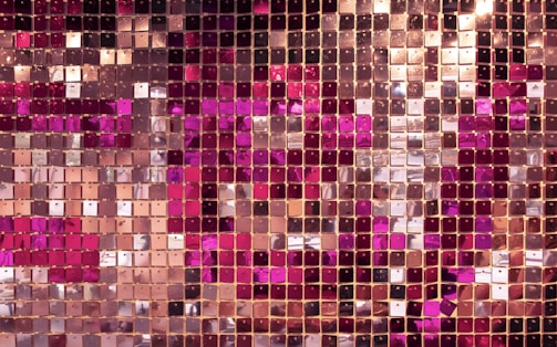 A close-up view of a shiny mosaic wall composed of numerous square panels. The panels are arranged in a grid pattern and vary in shades of pink, gold, and rose gold. The reflective surface of the panels creates a shimmering effect.