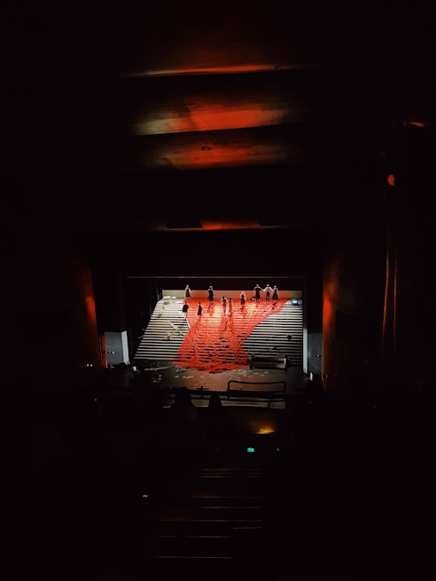 A dark theater setting with a stage illuminated by dramatic lighting. Several people are standing on the stage, with a large red fabric or pattern spread across the stairs. The audience area is dimly lit, emphasizing the focus on the stage.