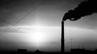A silhouette of an industrial chimney emitting dark smoke into the sky, with the sun partially visible through a hazy cloud cover. The scene is monochromatic, with strong contrasts between light and shadow.