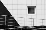 A modern architectural facade featuring geometric designs with a monochrome color scheme. A small rectangular window is set in a grid-patterned wall, with a diagonal black section contrasting against the predominant white. In the foreground, a metal railing runs diagonally across, adding a layer of depth and dimension.