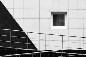 A modern architectural facade featuring geometric designs with a monochrome color scheme. A small rectangular window is set in a grid-patterned wall, with a diagonal black section contrasting against the predominant white. In the foreground, a metal railing runs diagonally across, adding a layer of depth and dimension.