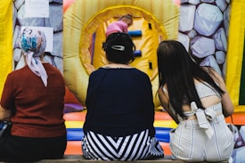 Three women are seated on a bench or platform, their backs facing the camera. They are observing an indoor inflatable play area with colorful obstacles. The walls have a stone-like pattern. A child, partially visible, is climbing in the play area.