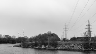 Power lines stretch across the open landscape beside a river, with a backdrop of industrial chimneys emitting smoke. Sparse vegetation and leafless trees are scattered along the riverbank, and the sky is overcast, contributing to the somber environment.
