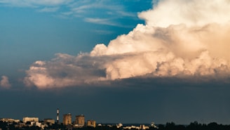 A large, towering cloud formation dominates the sky, with a mix of dark and light tones suggesting a dynamic weather change. Below, a cityscape with various buildings and a smokestack is visible against the horizon, partially obscured by the pending storm.