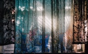 A window is adorned with semi-transparent curtains featuring a floral pattern. Sunlight filters through the fabric, creating intricate shadows and highlights on the surface. In the background, a vibrant blue fabric with floral designs is visible, adding a pop of color to the scene. The interplay of light and shadows gives the setting a dynamic and artistic feel.