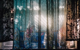 A window is adorned with semi-transparent curtains featuring a floral pattern. Sunlight filters through the fabric, creating intricate shadows and highlights on the surface. In the background, a vibrant blue fabric with floral designs is visible, adding a pop of color to the scene. The interplay of light and shadows gives the setting a dynamic and artistic feel.
