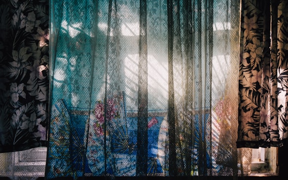 A window is adorned with semi-transparent curtains featuring a floral pattern. Sunlight filters through the fabric, creating intricate shadows and highlights on the surface. In the background, a vibrant blue fabric with floral designs is visible, adding a pop of color to the scene. The interplay of light and shadows gives the setting a dynamic and artistic feel.