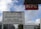 A multilingual sign indicating a border between sectors is displayed prominently in front of a modern urban backdrop. Next to it is a KFC sign. The sky is partly cloudy, and several buildings are visible in the background.