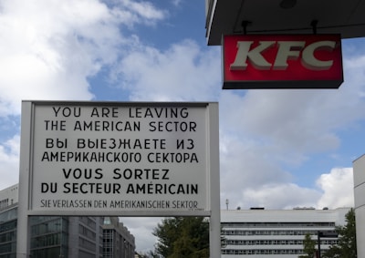 A multilingual sign indicating a border between sectors is displayed prominently in front of a modern urban backdrop. Next to it is a KFC sign. The sky is partly cloudy, and several buildings are visible in the background.
