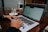 A person is typing on a laptop at a wooden table. A white coffee cup with a saucer and a sugar dispenser are nearby. The laptop screen displays a website or document in a window interface.