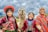 Three children dressed in vibrant, traditional Andean clothing are smiling broadly while standing next to a llama. Bright colors and intricate patterns of their garments create a lively and cheerful atmosphere against a backdrop of a cloudy sky and green fields.