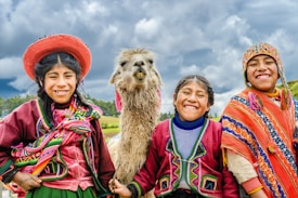 Three children dressed in vibrant, traditional Andean clothing are smiling broadly while standing next to a llama. Bright colors and intricate patterns of their garments create a lively and cheerful atmosphere against a backdrop of a cloudy sky and green fields.