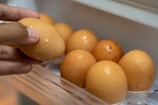 A hand is holding an egg, while other eggs are arranged in a plastic egg tray. The eggs are brown and appear fresh, with a slightly shiny surface.