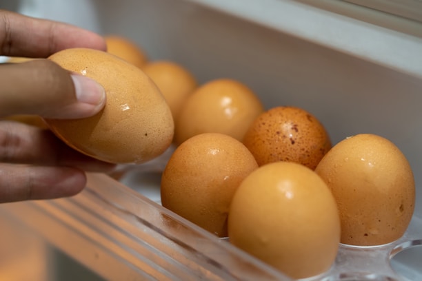 A hand is holding an egg, while other eggs are arranged in a plastic egg tray. The eggs are brown and appear fresh, with a slightly shiny surface.