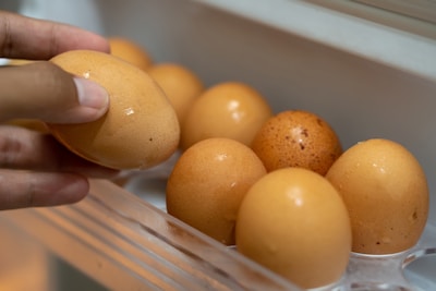 A hand is holding an egg, while other eggs are arranged in a plastic egg tray. The eggs are brown and appear fresh, with a slightly shiny surface.