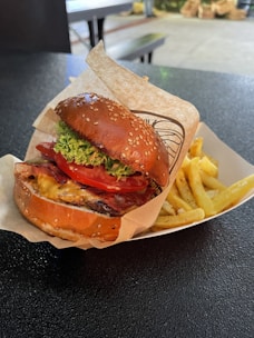 A well-arranged food scene featuring a sesame seed bun burger filled with crisp lettuce, fresh tomato slices, melted cheese, bacon, and a beef patty. The burger is placed on wrapping paper in a tray next to a serving of golden French fries. The setting appears to be a casual dining area with a textured dark tabletop.