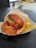 A well-arranged food scene featuring a sesame seed bun burger filled with crisp lettuce, fresh tomato slices, melted cheese, bacon, and a beef patty. The burger is placed on wrapping paper in a tray next to a serving of golden French fries. The setting appears to be a casual dining area with a textured dark tabletop.