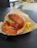 A well-arranged food scene featuring a sesame seed bun burger filled with crisp lettuce, fresh tomato slices, melted cheese, bacon, and a beef patty. The burger is placed on wrapping paper in a tray next to a serving of golden French fries. The setting appears to be a casual dining area with a textured dark tabletop.
