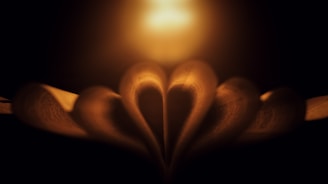Pages of a book are artistically folded to form a heart shape, illuminated by warm candlelight in a dark environment, creating a cozy and intimate atmosphere.