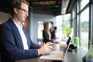 A person wearing glasses and a blue blazer is working on a laptop at a desk near large windows, with another person blurred in the background. The room is well-lit, with indoor plants and modern decor. A pair of JBL headphones and a green water bottle are placed on the desk.
