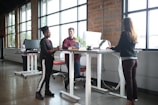 A modern office setting with three people interacting around standing desks. Large windows let in natural light, and there are computers and office supplies on the desks. The space has a casual and collaborative atmosphere.