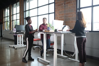 A modern office setting with three people interacting around standing desks. Large windows let in natural light, and there are computers and office supplies on the desks. The space has a casual and collaborative atmosphere.