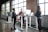 A modern office setting with three people interacting around standing desks. Large windows let in natural light, and there are computers and office supplies on the desks. The space has a casual and collaborative atmosphere.