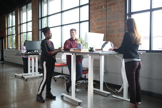 A modern office setting with three people interacting around standing desks. Large windows let in natural light, and there are computers and office supplies on the desks. The space has a casual and collaborative atmosphere.