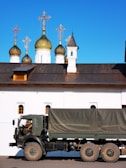 An army truck is parked in front of a white building with ornate green and gold domes topped with crosses. The architectural style is reminiscent of Russian Orthodox churches. A soldier is inside the truck.