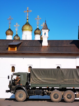 An army truck is parked in front of a white building with ornate green and gold domes topped with crosses. The architectural style is reminiscent of Russian Orthodox churches. A soldier is inside the truck.