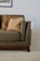 A comfortable gray and brown armchair partially visible on a soft beige carpet. The chair has textured upholstery and is accompanied by two beige pillows, one darker than the other. Soft light from the right creates gentle shadows, highlighting the cozy and inviting nature of the space. Above, a framed artwork is slightly visible.