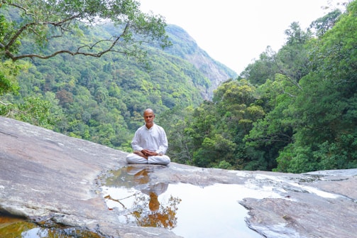 A person dressed in white sits peacefully in a meditative pose on a large rock surface with a small pool of water in front, surrounded by lush green forested mountains.