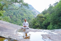 A person dressed in white sits peacefully in a meditative pose on a large rock surface with a small pool of water in front, surrounded by lush green forested mountains.