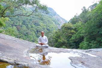 A person dressed in white sits peacefully in a meditative pose on a large rock surface with a small pool of water in front, surrounded by lush green forested mountains.