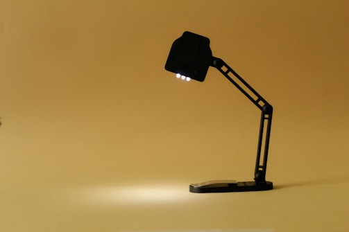 A black desk lamp with a modern, minimalist design is turned on, casting light on a plain beige background.