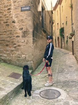 A person wearing sunglasses, a cap, and a tracksuit holds a red leash attached to a black dog. They stand in a narrow, cobblestone alley lined with stone buildings adorned with small windows and potted plants. A sign on the wall reads 'CUIDADO.' The atmosphere appears calm and historical with rustic architecture.