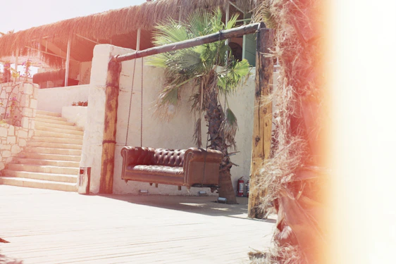 A sunlit patio featuring a rattan woven sofa set with colorful cushions, a metal hanging lantern glowing softly, and a sturdy umbrella base anchoring a large umbrella.