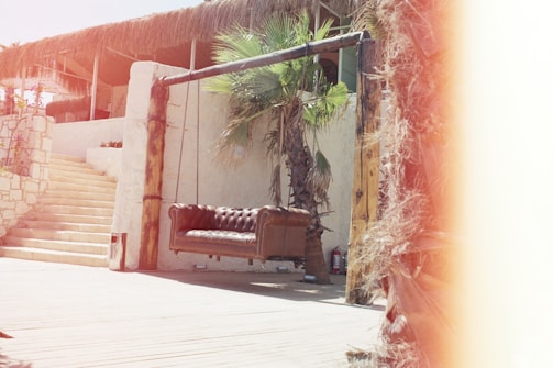 A sunlit outdoor patio with a rustic ambiance features a brown leather sofa hanging from a wooden frame. Stairs made of stone lead up to an area shaded by a thatched roof. A palm tree is present near the sofa, adding to the tropical feel.